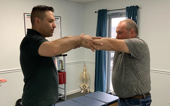 Two people standing in a chiropractic office performing an arm movement or range-of-motion exercise, with a treatment table, bookshelf, anatomical skeleton model, and window with blue curtains in the background