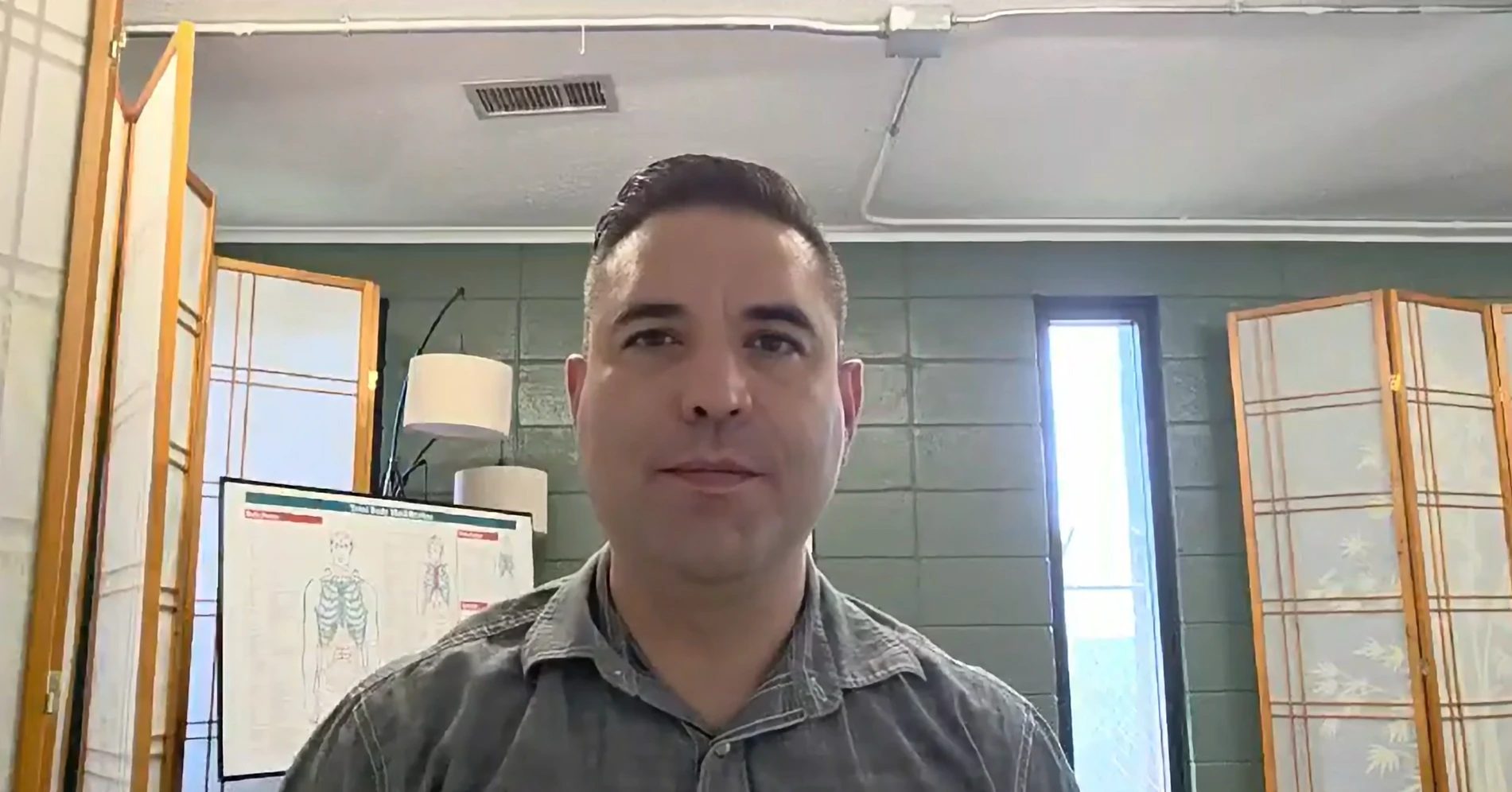 Dr Scott Forbes wearing a gray collared shirt seated indoors with a green cinder block wall in the background