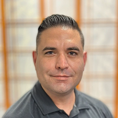 Dr Scott Forbes wearing a dark gray collared shirt, standing indoors in front of a softly focused background with wooden frames and light-colored panels